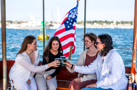 A group of four girls clinking their drinks together, while sitting in the back of the yacht Rum Runner for a Newport Scenic Sunset Cruise