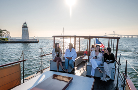 The stern of the Yacht Rum Runner II with a group of customers sitting enjoying drinks with a lighthouse in the background and the sun setting