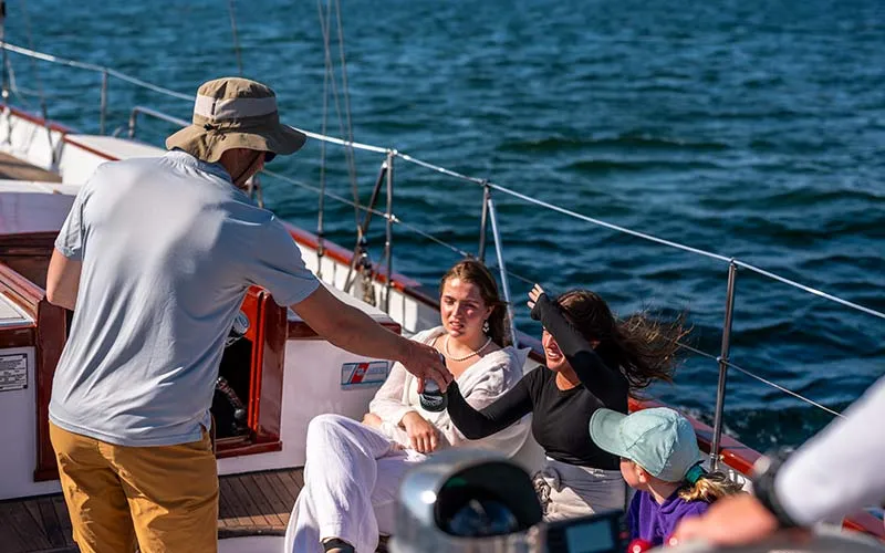 Newport_Classic-Afternoon-Sail-Madeleine_Slider-4 a group of people sitting on a sailboat, being handed drinks by a bartender