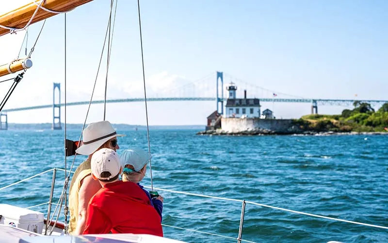 Newport_Classic-Afternoon-Sail-Madeleine_Slider-5 a couple looking out over a sailboat, towards a shore and a lighthouse in the distance