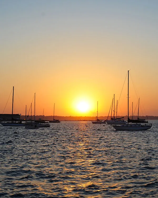 Newport_Private-Boat-Charters_Charter-Offerings-V3 a sunset over the horizon, with various different sailboats out on the water
