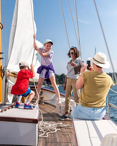 a woman taking a photo of two kids playing with the ropes of a sail